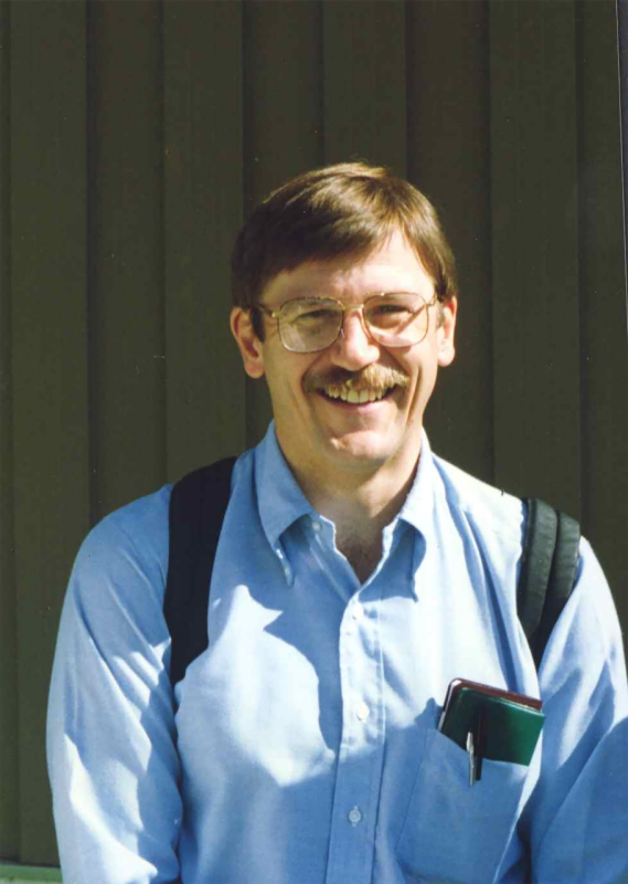 A man with large eye glasses and a pocket protector and blue shirt looks directly at the camera, smiling.
