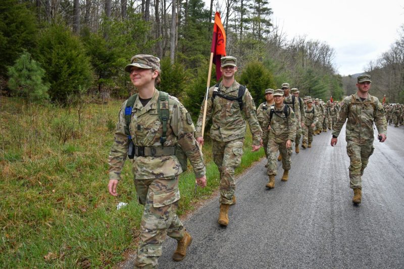 A young woman in full camouflage gear leads a  march of hundreds of cadets along a blacktop road.